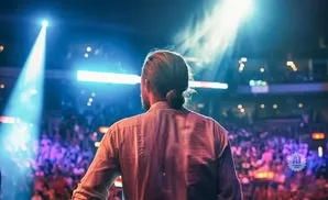 Man with hair in a bun on stage facing a blurred crowd under bright blue and white spotlights.