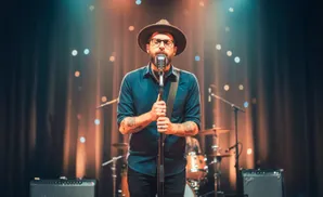 Man in a hat and glasses sings into a microphone on stage with a blurred background of lights and a drum set.