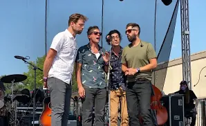 Four men in casual clothes sing into microphones on an outdoor stage.