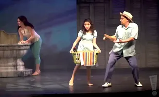 A man with a hat and glasses dances with a woman holding a laundry basket on stage.