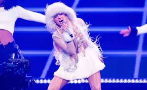A blonde singer in a white fur-trimmed outfit and a fluffy hat sings into a microphone on a blue, lit stage.