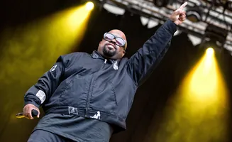 A man with sunglasses points his finger up on stage with yellow spotlights behind him.