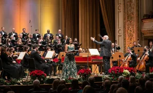 A female vocalist in a teal gown sings with a choir and orchestra performing in a concert hall.