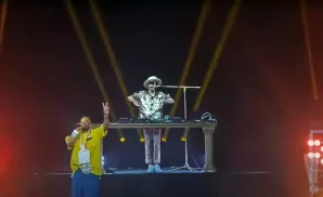 A rapper in a yellow shirt and large chain holds a microphone, while a DJ in a sequined shirt and hat spins records.