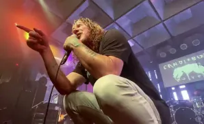 A male singer with long blonde hair crouches and sings into a microphone, pointing his finger towards the left.