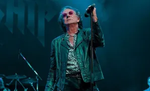 Man in a leather jacket and leopard print shirt sings into a microphone on stage.