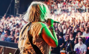 Singer on stage with colorful hair, holding a microphone, with a blurred crowd in the background.