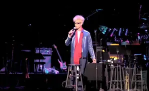 A man with blond hair and glasses, wearing a red scarf and blue jacket, holds a microphone on stage.