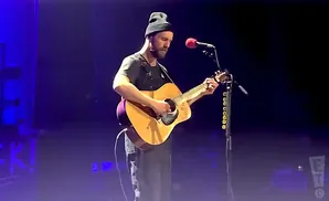 A man in a beanie plays an acoustic guitar on stage, illuminated by blue and purple lights.
