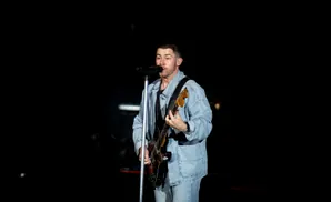 Nick Jonas performs on stage with a guitar and microphone, wearing a denim jacket and jeans.