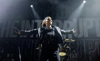 Singer in a star-print shirt and leather jacket on stage, holding a microphone, with 