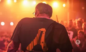 Man in a black t-shirt with an orange graphic stands in front of a crowd under bright orange lights.