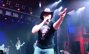 A man in a cowboy hat sings into a microphone on stage, wearing a t-shirt with an American flag design.