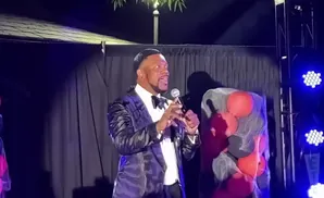 Man in a sparkly suit speaks into a microphone on a stage with blue lights and a balloon display.