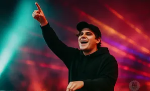 Man in a black hoodie and baseball cap points up with his right hand while smiling at a concert with colorful lights.