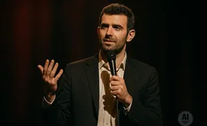 Comedian on stage holding a microphone and gesturing with his hand.