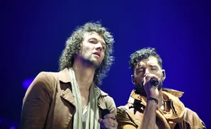 Two men singing into microphones on a stage with a blue background.