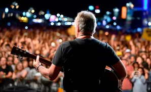 Back view of a guitarist playing for a large, blurred crowd at a concert with bright stage lights.