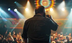 A man with a beard sings into a microphone on stage, with a crowd and spotlights behind him.