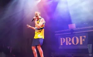 Rapper PROFF performs on stage in a yellow sports jersey and dark shorts, holding a microphone.