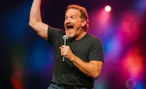 A man with an open mouth and raised arm holds a microphone on a stage with colorful lights.