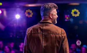 Man in a brown jacket seen from behind on a stage with purple lights and a blurred audience.