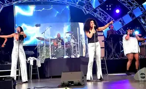 A disco band performs on stage, with a female singer in a sparkly jumpsuit in the foreground.