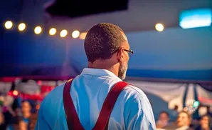 Man with glasses and red suspenders faces away from camera on a stage with blurred audience.