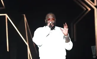 Rapper Rick Ross performs on stage in a white sweatshirt and sunglasses, holding a microphone.