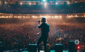 Singer with long hair on stage in front of a large, illuminated crowd at a concert.