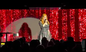 A drag performer with long blonde hair wears a sparkly green dress and sings into a microphone on a red glittery stage.