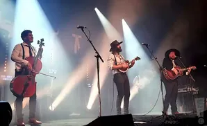 Three musicians perform on stage: a cellist, a mandolin player, and a guitarist.