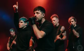 A band performs on stage with red and blue lighting. The lead singer is in the foreground singing into a microphone.