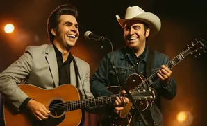 Two men playing guitars and singing on stage. One wears a grey suit, the other a denim jacket and cowboy hat.