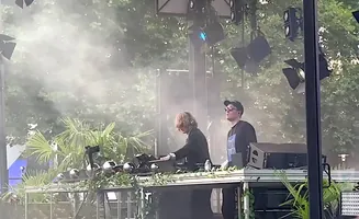 Two DJs on a stage with smoke and palm trees.