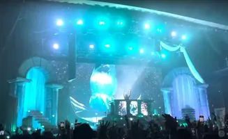A DJ performs on a stage with a large screen displaying an icy landscape, while an audience cheers.