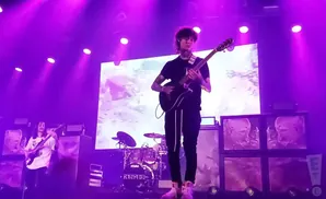A guitarist performs on a stage with purple lighting, playing a black electric guitar.