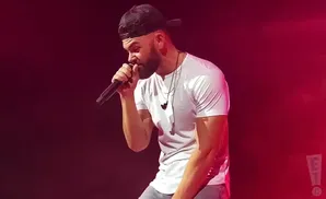 A man in a white t-shirt and baseball cap sings into a microphone on a stage with red lighting.