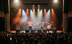 Two men stand on a dimly lit stage, with a crowd in the foreground. The man on the left wears a pink shirt, the man on the right a plaid shirt and cap.