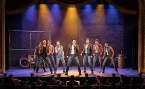 A male musical ensemble performs on stage with brick and chain-link fence backdrop.