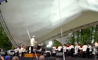 An orchestra performs under a white canopy. A conductor in a light-colored jacket leads the musicians.