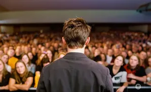 Man in a black jacket faces a large, blurred audience in an indoor venue.