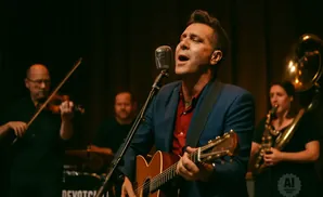 A singer in a blue jacket and red shirt plays an acoustic guitar while a violinist and saxophonist perform behind him.