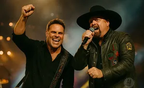 Two men on stage, one raising his fist, the other singing into a microphone, both wearing black shirts and jackets.