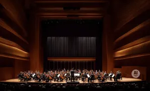 An orchestra performs on stage in a concert hall with warm, wood-paneled walls.
