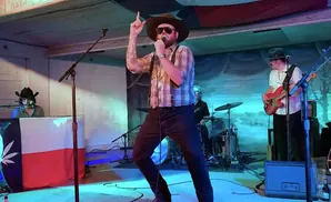 A band performs on stage, with a singer in a hat and plaid shirt in the foreground.