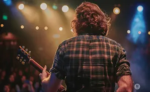 A guitarist with long, curly hair plays on a stage lit by warm stage lights.