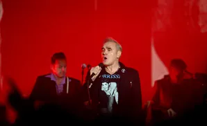 Singer Morrissey on stage in front of a red backdrop, wearing a black jacket over a t-shirt with his face on it.