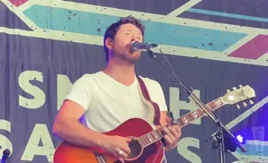 A man in a white t-shirt plays a red acoustic guitar on stage, singing into a microphone.