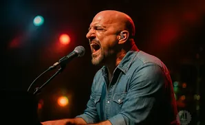Bald man singing intensely into a microphone on stage with colorful lights.